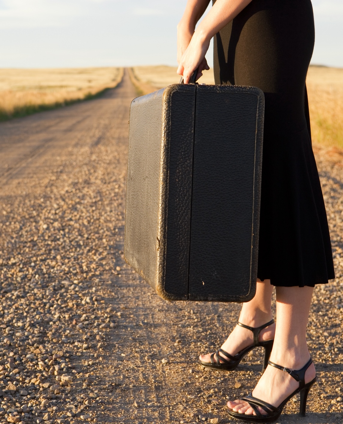 Woman in a black dress and heels waiting along side a dirt road with her suitcase.